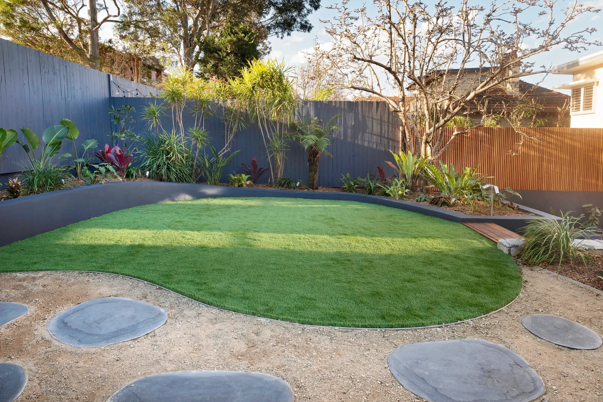 Greenwich rear garden with artificial turf, curved rendered planter wall and tropical planting