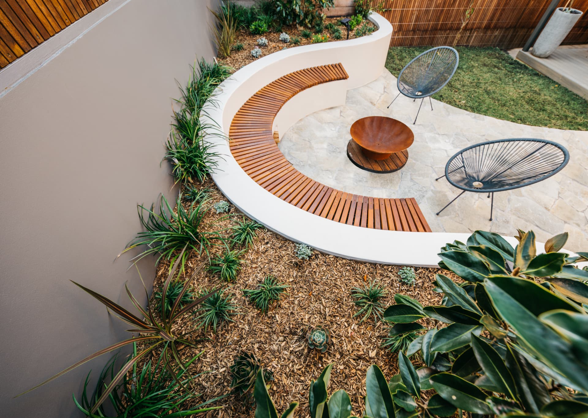 Elevated view of the curved rendered wall, timber bench and stone paved fire pit court at Wareemba