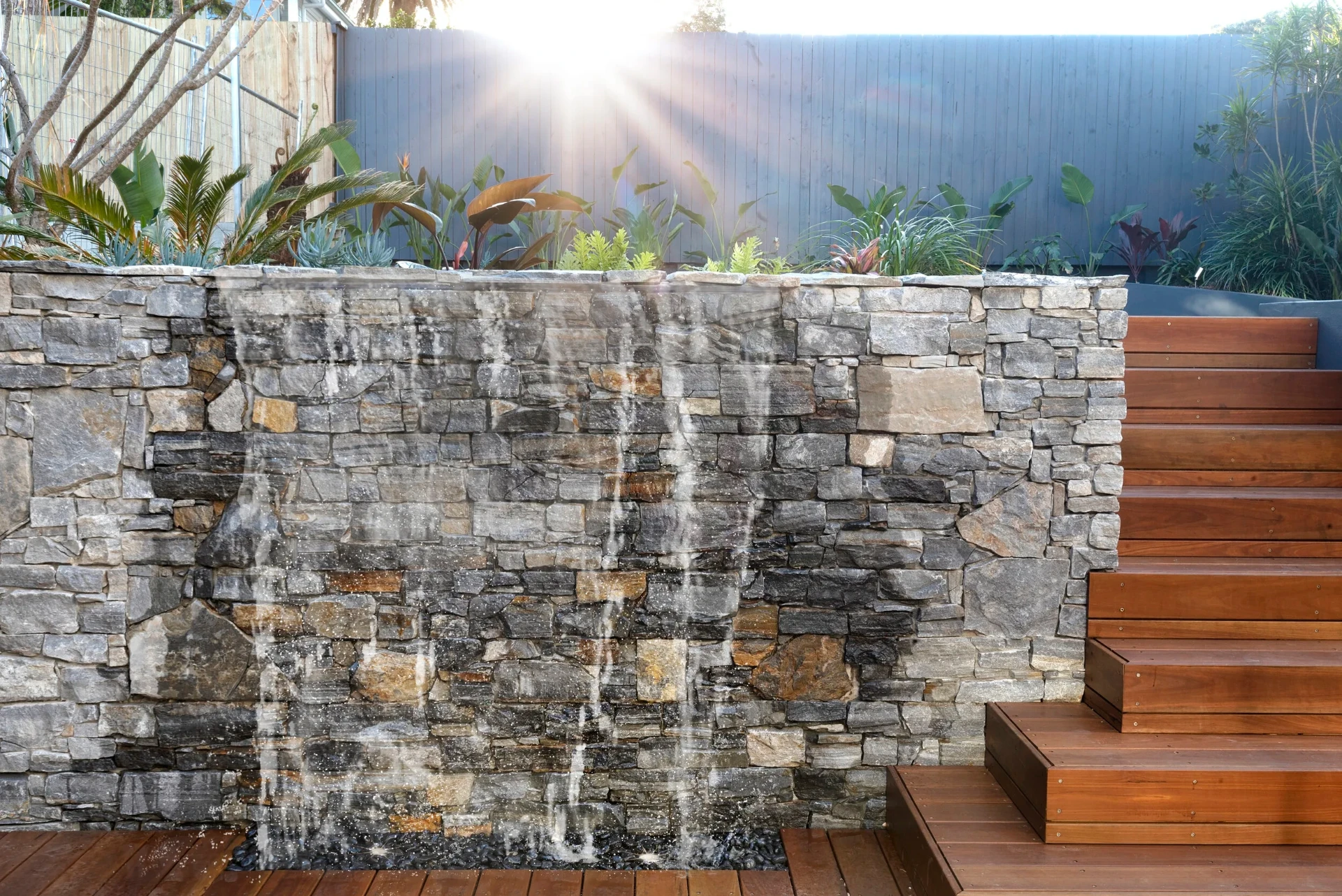 Stone-clad water feature beside hardwood stairs and layered planting in the Greenwich garden