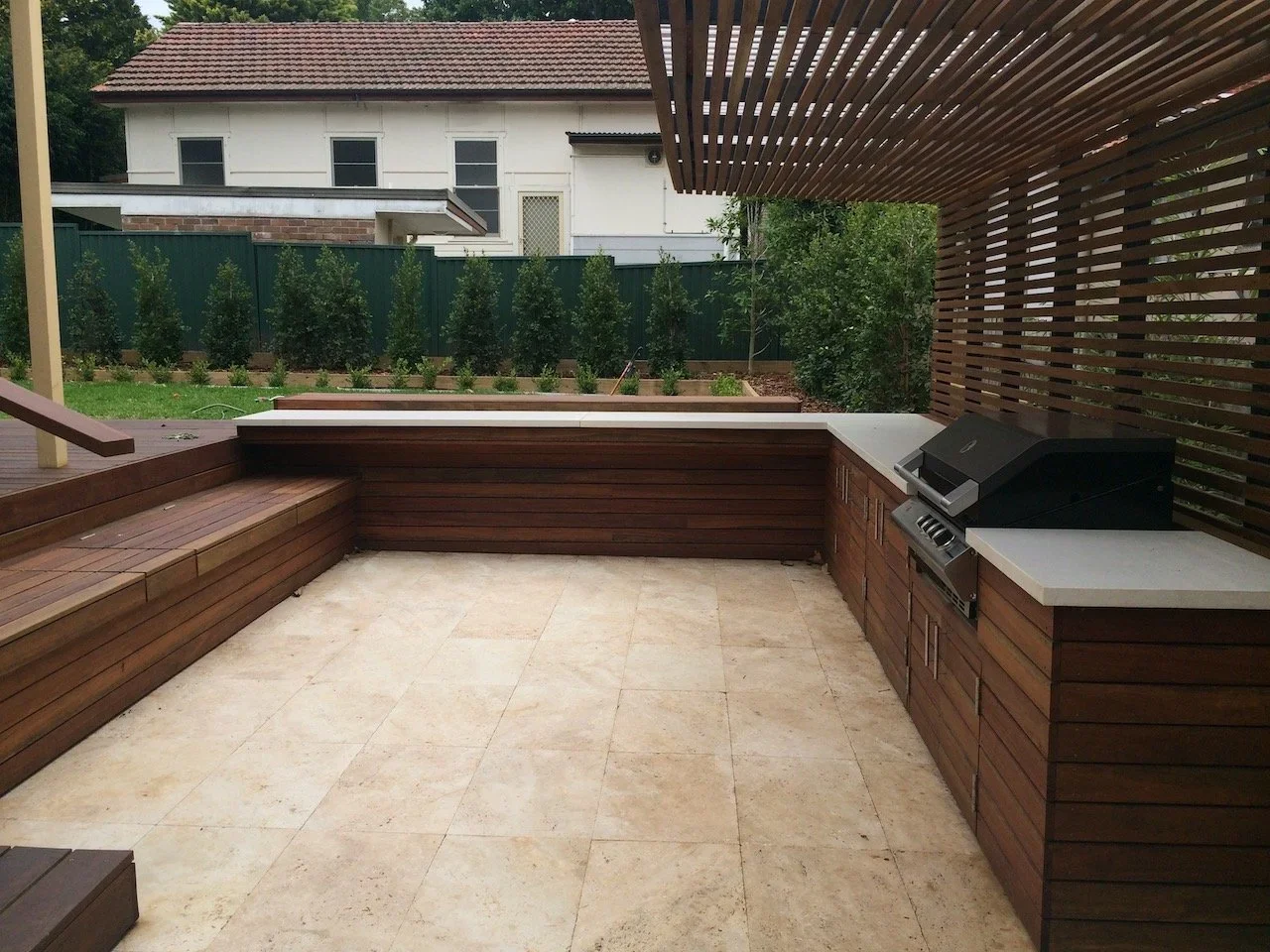 Outdoor kitchen and built-in bench seating beside travertine paving and a screened boundary
