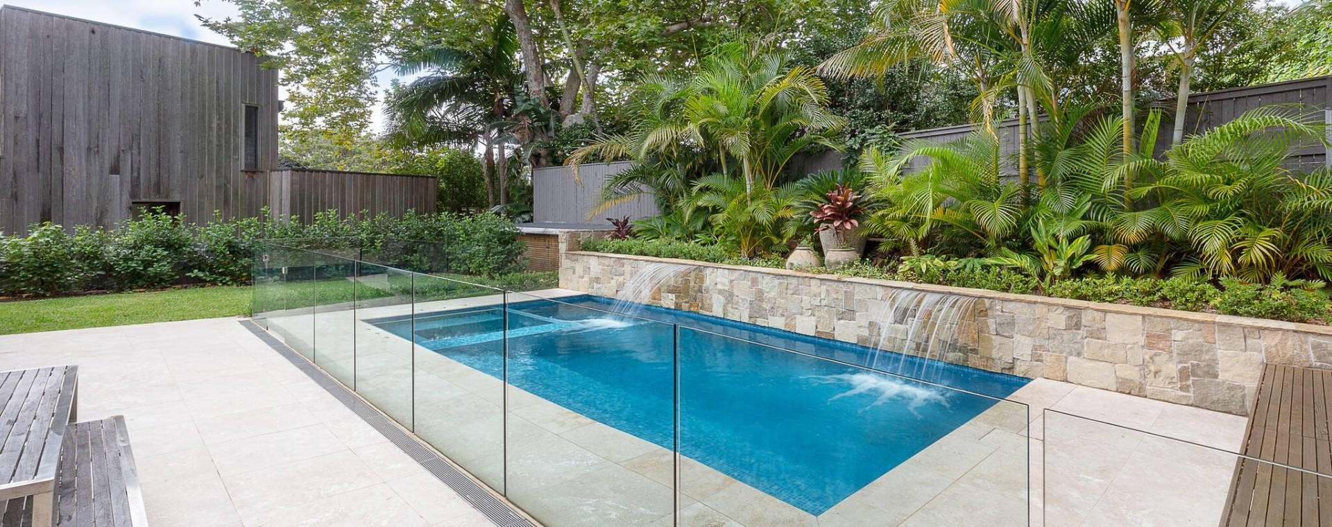 Tiled pool with sandstone waterfall wall and frameless glass fence