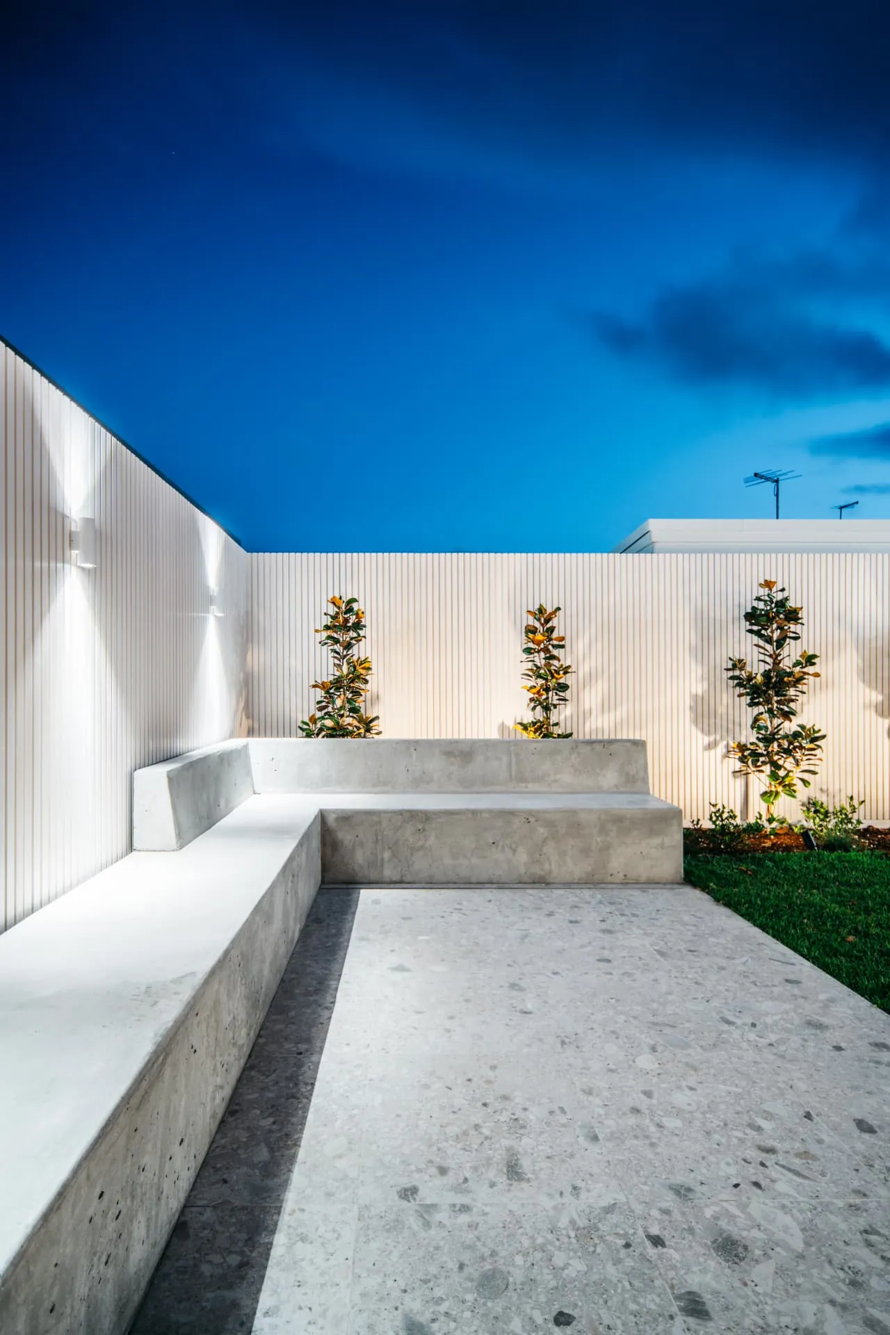 L-shaped board-formed concrete bench at dusk with uplit battened fence and magnolias